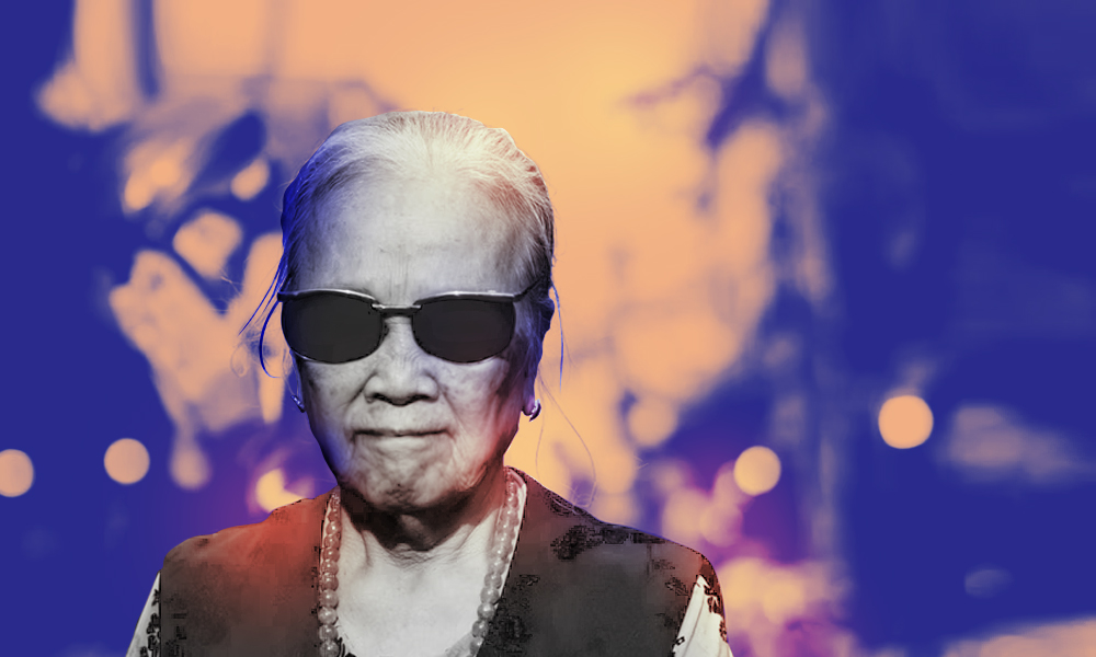 A black-and-white image of an older woman from the shoulders up. She has white hair pulled back behind her head and, and she is wearing sunglasses, pearls, and a black dress.