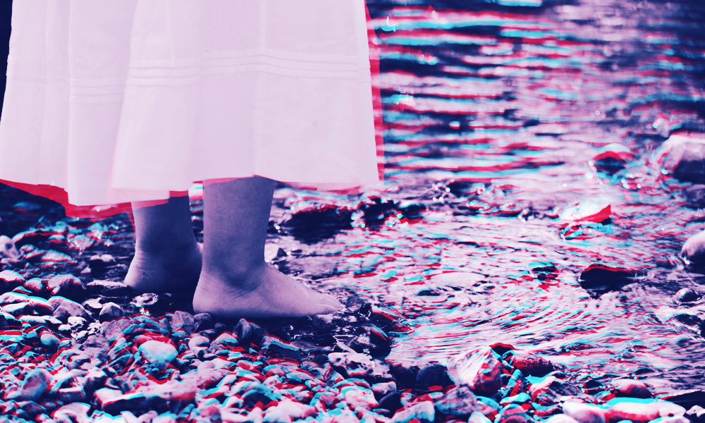 A close-up purple-and-pink image of a child's feet standing atop small rocks at the edge of a river.