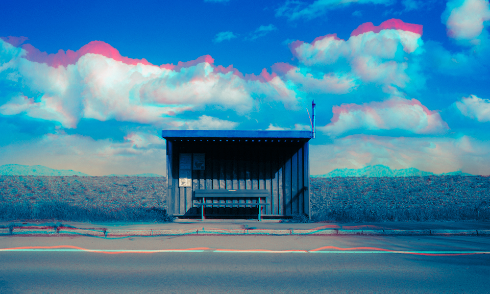 An empty blue bus stop shelter with a bench in front of a big field and a blue sky with white and pink clouds.